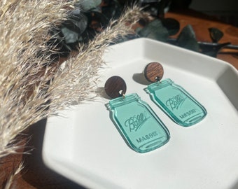 May include: A pair of teal blue glass mason jar earrings with wooden tops. The jars have the words "Ball Mason" etched on them.
