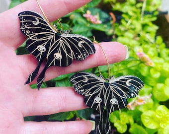 Könnte beinhalten: Ein Paar schwarze Creolen mit einem goldfarbenen Ring. Jeder Ohrring zeigt eine schwarze Motte mit goldenen Details, darunter Sterne, Monde und Augen.