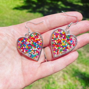 May include: A pair of silver heart-shaped earrings with colorful sprinkles embedded in clear resin.