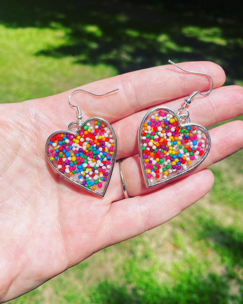 May include: A pair of silver heart-shaped earrings with colorful sprinkles embedded in clear resin.