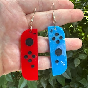 Puede incluir: Pendientes con diseños de mandos de videojuegos, uno rojo y otro azul. El pendiente rojo tiene botones negros y un círculo negro, el azul tiene botones negros y un círculo negro. Ambos tienen ganchos plateados.