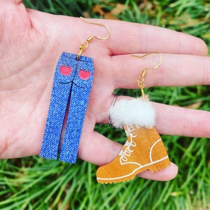 May include: A pair of earrings featuring a blue denim jean and a brown boot with white fur trim. The jean has two red hearts on the back pockets. The earrings are made of wood and have gold hooks.