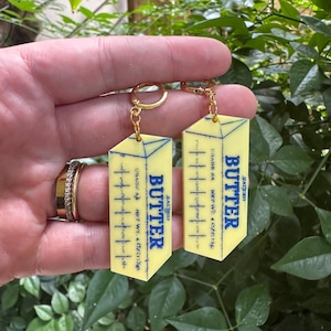 May include: A pair of novelty earrings featuring a yellow butter stick design with the word "BUTTER" in blue. The earrings have gold-colored hoops and are held against a backdrop of green foliage. The text "GRADE AA NET WT. 4.020156" is visible.