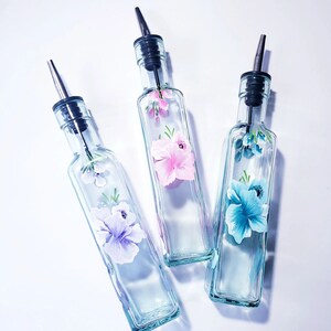 Puede incluir: Tres botellas de aceite de oliva de vidrio con diseños florales. Las botellas son de vidrio transparente con un pico de vertido negro y un diseño floral pintado en la parte delantera. Las flores son rosas, moradas y azules.