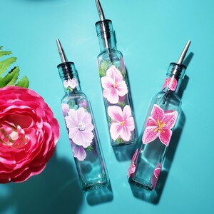 Puede incluir: Tres botellas de vidrio transparente con boquillas de acero inoxidable, cada una pintada a mano con un diseño floral rosa diferente. Las botellas están dispuestas sobre una superficie turquesa brillante, con una gran flor rosa.