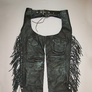May include: Black leather chaps with fringe detail and a belt. The chaps have a pocket on the right leg.