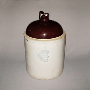 May include: A brown glazed stoneware crock with a white body. The crock has a number "5" and a blue leaf design on the front.