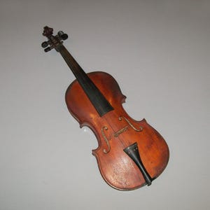 May include: A vintage violin with a worn brown finish and black strings. The violin has a simple design with a single f-hole and a black chin rest.