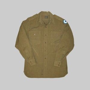 Puede incluir: Una camisa de trabajo de manga larga de color verde oliva con dos bolsillos en el pecho y cuello abotonado. La camisa tiene un parche en el hombro derecho con un diseño azul y blanco.