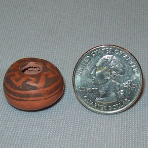Può includere: Un piccolo vaso di terracotta marrone con disegni geometrici neri. Il vaso è circa delle dimensioni di una moneta da 25 centesimi di euro.