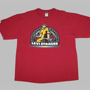 Op de afbeelding: Rood T-shirt met korte mouwen met een afbeelding van een arbeider die een hamer vasthoudt voor een tandwiel met de tekst "Levi Strauss & Co."