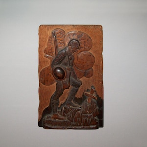 May include: A wooden relief carving of a soldier carrying a rifle and a helmet, standing on a hill with a cross in the background.