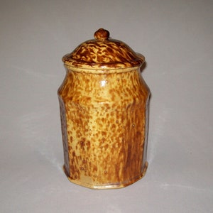 May include: A yellow and brown glazed ceramic jar with a lid. The jar has a rounded body and a slightly flared top. The lid has a small knob on top.