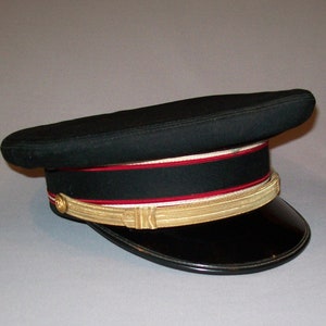 Puede incluir: Un sombrero negro de estilo militar con una banda dorada y ribetes rojos y blancos.