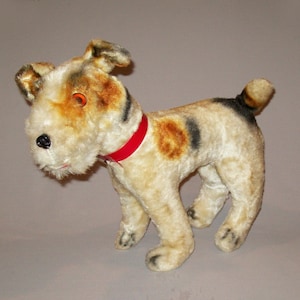 Può includere: Un cane di peluche vintage con pelliccia marrone, bianca e nera. Il cane ha un collare rosso e un occhio arancione.