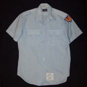 May include: A light blue short-sleeved shirt with a button-up front and two flap pockets. The shirt has a small patch on the right sleeve with a gold eagle design. The shirt is labeled "Cadet" and "Made in U.S.A."