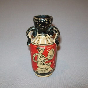 May include: A small black and red ceramic vase with two handles and a white and black design of a woman playing a lyre.