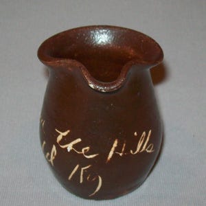 May include: A small, brown ceramic pitcher with a spout and a rounded body. The pitcher has white handwritten text that reads "the Hills 15". The pitcher is likely handmade.