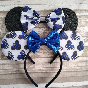 May include: Black headband with two pairs of mouse ears. The ears are covered in white fabric with a blue and black thin striped pattern and a small blue and black Mickey Mouse silhouette. One pair of ears has a blue sequin bow and the other has a white bow with silver glitter.