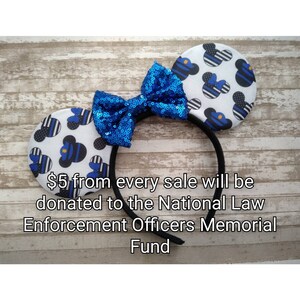 May include: Blue and white mouse ears headband with a blue sequined bow. The headband has a thin black band and the ears are printed with a pattern of blue and white stripes and a small blue and white police officer figure. The text "$5 from every sale will be donated to the National Law Enforcement Officers Memorial Fund" is printed below the headband.