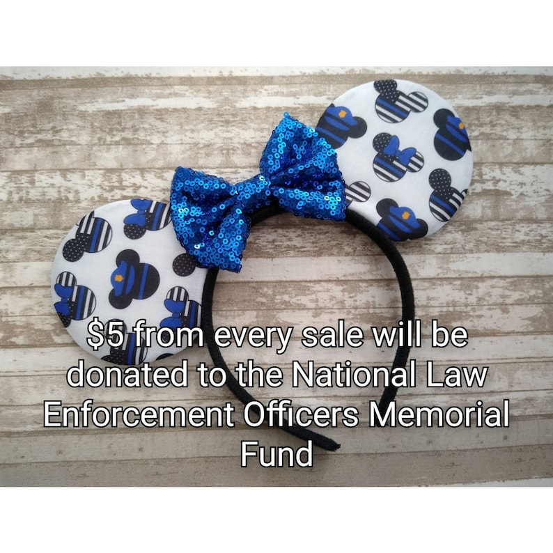 May include: Blue and white mouse ears headband with a blue sequined bow. The headband has a thin black band and the ears are printed with a pattern of blue and white stripes and a small blue and white police officer figure. The text "$5 from every sale will be donated to the National Law Enforcement Officers Memorial Fund" is printed below the headband.
