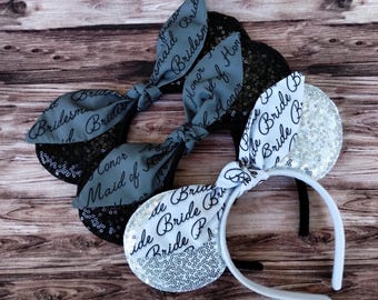 May include: Three headbands with sequined ears and fabric bows. Two headbands have black ears and gray bows with the words "Bridesmaid" and "Honor" printed on them. The third headband has white ears and a white bow with the word "Bride" printed on it.