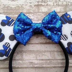 May include: A pair of white mouse ears with a blue and black thin stripe pattern featuring a blue police officer Mickey Mouse silhouette. The ears are attached to a black headband with a large blue sparkly bow.