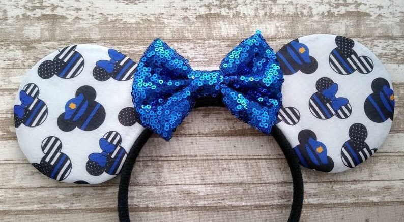 May include: A pair of white mouse ears with a blue and black thin stripe pattern featuring a blue police officer Mickey Mouse silhouette. The ears are attached to a black headband with a large blue sparkly bow.