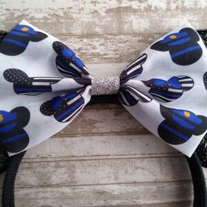May include: Black and white mouse ears headband with a blue and black bow featuring a thin blue line pattern and a silver glitter center. The bow is decorated with a repeated pattern of a cartoon mouse head wearing a police officer's hat.