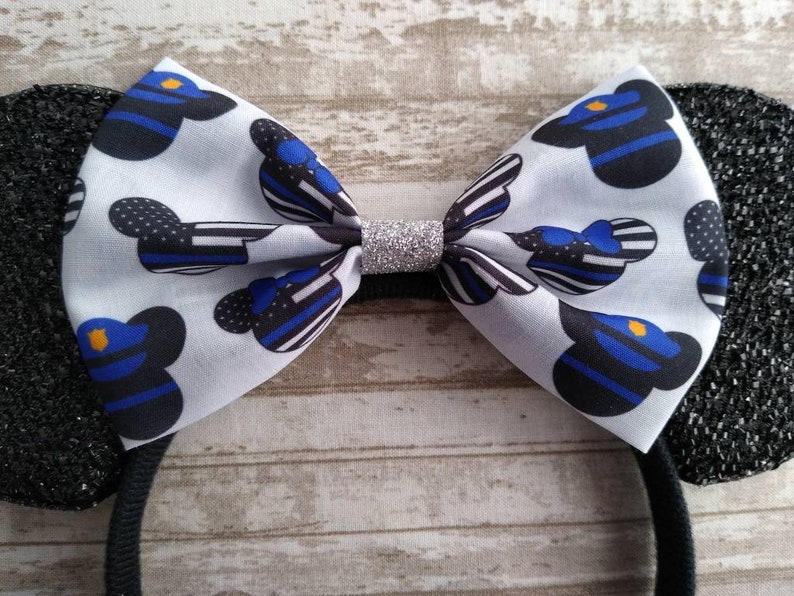 May include: Black and white mouse ears headband with a blue and black bow featuring a thin blue line pattern and a silver glitter center. The bow is decorated with a repeated pattern of a cartoon mouse head wearing a police officer's hat.