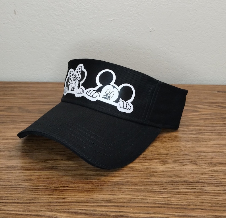 May include: A black visor with a white Mickey Mouse and Minnie Mouse design. The characters are peeking over the brim of the visor.