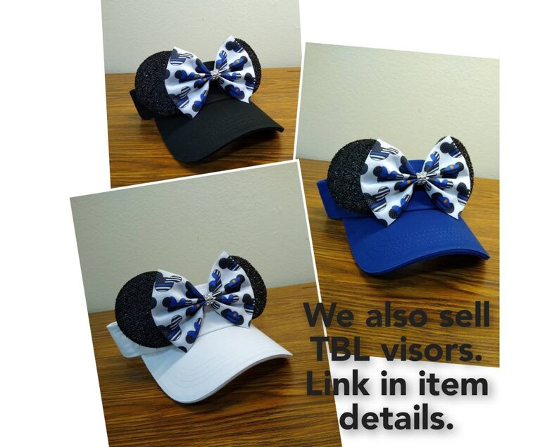 May include: Three different colored visors with a black and blue bow with a rhinestone in the center. The bow is shaped like Mickey Mouse ears. The text "We also sell TBL visors. Link in item details." is at the bottom of the image.