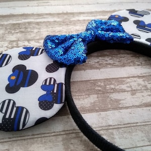 May include: Blue and black mouse ears headband with a blue sequined bow. The ears are decorated with a white background and a pattern of blue and black thin stripes and a black silhouette of a mouse head with a blue police officer's hat.