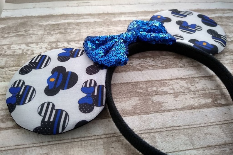 May include: Blue and black mouse ears headband with a blue sequined bow. The ears are decorated with a white background and a pattern of blue and black thin stripes and a black silhouette of a mouse head with a blue police officer's hat.