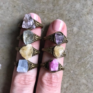 Puede incluir: Conjunto de seis anillos con diferentes piedras de cristal natural de colores engastadas en monturas de metal de tono dorado antiguo. Las piedras incluyen amatista, aventurina verde, citrino, selenita y cuarzo rosa.