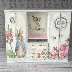 Könnte beinhalten: Ein weißer Miniatur-Kleiderschrank aus Holz mit Peter-Hase-Illustrationen, einem Kaninchen in blauer Jacke, einem Vogel auf einer Gießkanne und Blumenmotiven. Die rechte Tür zeigt ein kreisförmiges Logo mit dem Text Peter Rabbit und kleine Goldknöpfe.