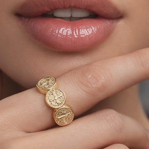 close-up beauty photo of a woman with soft, sensual glossy lips, natural glowing skin, and manicured hand gently touching her face, showcasing a gold medallion statement ring; elegant, minimal, luxury jewelry product image designed to highlight texture and shine.