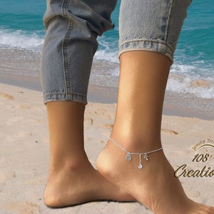 Puede incluir: Un brazalete de tobillo plateado con varios colgantes, incluyendo un círculo, una estrella y una luna creciente. El brazalete se lleva en el tobillo de una persona en una playa de arena.
