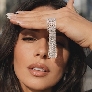 May include: A silver-toned statement ring with a rectangular top and dangling chain fringe. The ring features intricate detailing and is held up to the face. The person is wearing a black blazer.