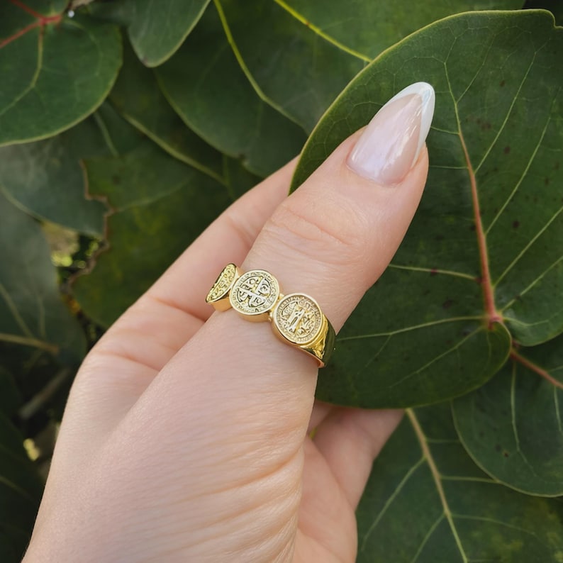 Puede incluir: Un anillo dorado con tres medallones circulares en relieve. El anillo, usado en un dedo, destaca sobre hojas verdes. El dise&ntilde;o incluye s&iacute;mbolos religiosos. El anillo probablemente est&aacute; hecho de metal dorado.