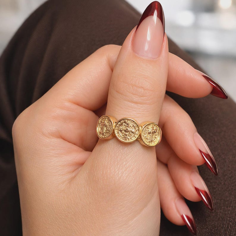 Close-up of a woman’s hand wearing a gold triple coin ring, shown on smooth, well-groomed fingers with glossy almond-shaped ombré red manicure. The hand rests on dark brown fabric under soft professional studio lighting, highlighting the detailed medallion design and polished finish of the ring.
