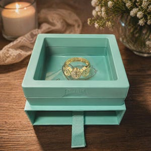 Puede incluir: Un anillo dorado con tres medallones decorativos, exhibido en una caja azul claro. El anillo est&aacute; centrado en la caja, que tiene un caj&oacute;n extra&iacute;ble. La caja est&aacute; sobre una superficie de madera, con una vela encendida y flores en el fondo.