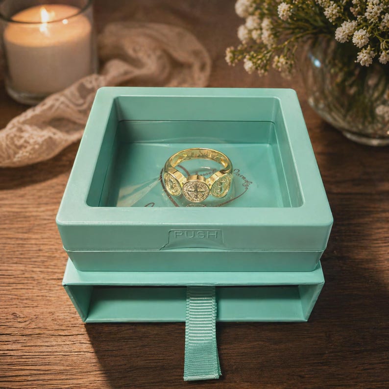 Puede incluir: Un anillo dorado con tres medallones decorativos, exhibido en una caja azul claro. El anillo est&aacute; centrado en la caja, que tiene un caj&oacute;n extra&iacute;ble. La caja est&aacute; sobre una superficie de madera, con una vela encendida y flores en el fondo.