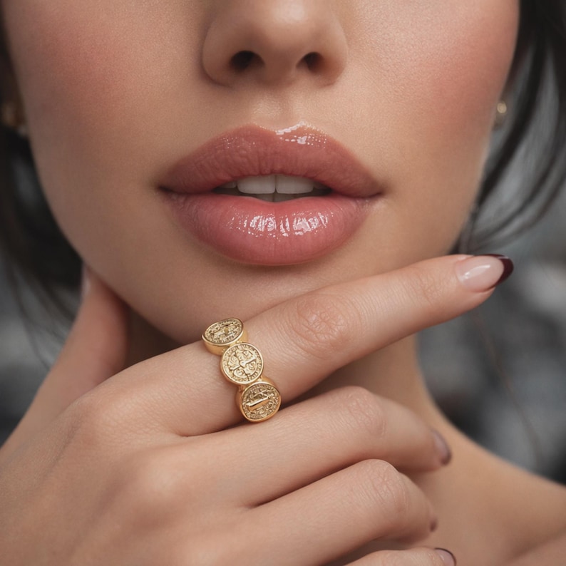 close-up beauty photo of a woman with soft, sensual glossy lips, natural glowing skin, and manicured hand gently touching her face, showcasing a gold medallion statement ring; elegant, minimal, luxury jewelry product image designed to highlight texture and shine.
