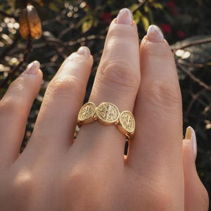 Puede incluir: Anillo dorado con tres medallones circulares con grabados detallados. El anillo se lleva en un dedo, mostrando su dise&ntilde;o y artesan&iacute;a. El anillo tiene un peque&ntilde;o dije colgante.