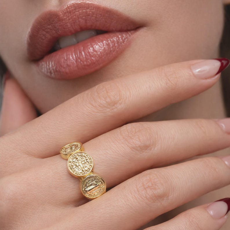 Gold Saint Benedict ring with three round medallions on a woman’s hand, close-up studio shot showing detailed engravings, polished finish, natural skin tone, elegant manicure, and soft neutral background.