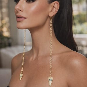 May include: Long gold chain earrings with a heart-shaped pendant featuring an eye design. The earrings are made of gold-colored metal and hang down from the ear. The earrings are a stylish accessory.