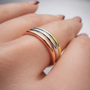 May include: A close-up of three rings in rose gold, silver, and yellow gold. The rings are stacked together on a finger, showcasing their different metallic colors and smooth, rounded bands. The rings are simple and elegant.
