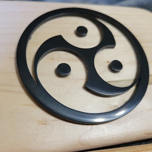 Pu&ograve; includere: Un simbolo triquetra nero, noto anche come nodo triplo, &egrave; intagliato su una superficie in legno chiaro.