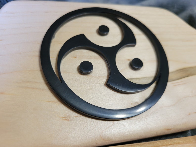 Pu&ograve; includere: Un simbolo triquetra nero, noto anche come nodo triplo, &egrave; intagliato su una superficie in legno chiaro.